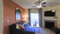 This single-floor residence in Euless, Texas, offers a warm and inviting living environment designed for comfortable everyday living. The living room serves as the heart of the space, showcasing a bold accent wall in a rich terracotta hue that adds a vibrant, artistic touch to the room. A large piece of wall art complements the color palette, enhancing the room's character. Plush carpeted flooring runs throughout, adding warmth underfoot. A ceiling fan with integrated lighting provides both comfort and illumination, while a wall-mounted flat-screen television offers modern entertainment convenience. The living area features a classic fireplace with a clean white mantle, serving as an elegant focal point. A sliding glass door, dressed with floor-to-ceiling draping curtains, floods the space with natural light and opens to a private balcony, offering a seamless connection to the outdoors. The overall ambiance is cozy yet stylish, blending warm tones with functional design elements that make this Euless residence a welcoming place to call home.