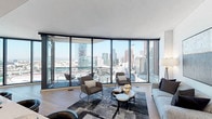 This single-floor high-rise residence is defined by its dramatic floor-to-ceiling glass walls, which frame expansive panoramic views of a major city skyline and surrounding urban landscape from nearly every room. Light wood flooring flows consistently throughout, complementing the clean, contemporary aesthetic that unifies the entire space.

The main living area is anchored by a curved glass curtain wall, flooding the room with natural light and offering sweeping cityscape views. Adjacent to it, a dedicated dining nook sits within a glass corner, fitted with a modern pendant chandelier and direct balcony access. The kitchen is sleek and well-appointed, featuring high-gloss cabinetry, stainless steel appliances including a professional-grade range and refrigerator, and a generous island with bar seating finished with gold-accented stools.

The primary bedroom enjoys large picture windows with broad city views, while a walk-in closet offers custom built-in shelving. A secondary living or sitting room provides a quieter retreat. The primary bathroom features a glass-enclosed walk-in shower with mosaic tile, a double vanity, and clean white finishes, while a secondary bathroom includes a soaking tub with marble-style surround tiling and a lit vanity mirror.