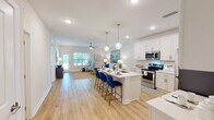 Located at 7155 N Blue Angel Pkwy in Pensacola, Florida, this single-story residence offers a bright, modern interior with a cohesive coastal-inspired design. The open-concept layout seamlessly connects the kitchen, dining, and living areas, creating a spacious and functional flow throughout. The kitchen is outfitted with white shaker cabinetry, stainless steel appliances, a large center island with seating for multiple bar-stool chairs, pendant lighting, and light-toned hardwood-style flooring that continues throughout the main living areas. The living room benefits from abundant natural light through sliding glass doors and windows, complemented by a ceiling fan overhead. Two well-appointed bedrooms each feature ceiling fans, ample natural light from windows with views of the surrounding area, and plush carpeted flooring. The primary bathroom includes a double-sink vanity with white cabinetry, a large mirror, and a tub-shower combination. A secondary full bathroom offers a similarly clean, white vanity with modern fixtures. A dedicated laundry area houses a full-size washer and dryer. Throughout the residence, neutral tones and clean white walls contribute to a bright, airy ambiance well-suited to the Florida climate.