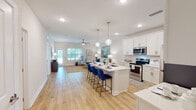 Located at 7155 N Blue Angel Pkwy in Pensacola, Florida, this single-story residence offers a bright, modern interior with a cohesive coastal-inspired design. The open-concept layout seamlessly connects the kitchen, dining, and living areas, creating a spacious and functional flow throughout. The kitchen is outfitted with white shaker cabinetry, stainless steel appliances, a large center island with seating for multiple bar-stool chairs, pendant lighting, and light-toned hardwood-style flooring that continues throughout the main living areas. The living room benefits from abundant natural light through sliding glass doors and windows, complemented by a ceiling fan overhead. Two well-appointed bedrooms each feature ceiling fans, ample natural light from windows with views of the surrounding area, and plush carpeted flooring. The primary bathroom includes a double-sink vanity with white cabinetry, a large mirror, and a tub-shower combination. A secondary full bathroom offers a similarly clean, white vanity with modern fixtures. A dedicated laundry area houses a full-size washer and dryer. Throughout the residence, neutral tones and clean white walls contribute to a bright, airy ambiance well-suited to the Florida climate.