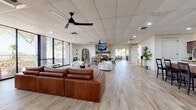 This expansive single-floor space is thoughtfully designed with a modern yet warm aesthetic, combining functionality with inviting interiors throughout. Light wood-look flooring runs continuously across the entire layout, unifying the open-plan design and contributing to a bright, airy atmosphere.

The main gathering area is generously proportioned, anchored by a substantial leather sofa and complemented by white accent chairs arranged around a central seating zone. Dual ceiling fans and recessed lighting ensure consistent comfort and illumination throughout. A notable stone-clad fireplace serves as a focal point along one wall, with a wall-mounted television above, creating a cozy yet stylish lounge environment.

Large floor-to-ceiling windows and glass doors along one side of the space flood the interior with natural light while providing clear views of the surrounding outdoor landscape. A dedicated dining area with a round table and chairs sits adjacent to the fireplace, ideal for smaller gatherings.

The fully equipped kitchen features rich dark wood cabinetry, marble-look countertops, stainless steel appliances, and a central island with bar seating. An arched display cabinet adds a decorative architectural touch nearby.

An accessible bathroom with grab bars, a glass vessel sink, and tasteful botanical artwork completes the space. Throughout, the design balances comfort, accessibility, and contemporary style.