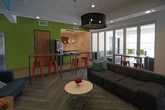 This single-floor communal space is thoughtfully designed to encourage social interaction, offering multiple distinct zones within an open, connected layout. The main lounge area is anchored by a large, plush sectional sofa set atop herringbone-patterned carpet, paired with a round coffee table and illuminated by a striking oversized drum pendant light. Bold teal accent walls define a cozy media nook, complete with a wall-mounted flat-screen TV, a low wood media console, and eclectic seating including a retro-style armchair and rustic log stools. Vibrant green accent walls energize the adjacent kitchen and dining area, which features high-top bar tables and colorful metal stools. The kitchenette is well-appointed with warm wood cabinetry, stainless steel appliances including a built-in oven and under-counter refrigerator, a utility sink, and a herringbone tile backsplash. A coffee station adds to the functionality of this area. Large glass bi-fold doors connect the interior seamlessly to an outdoor patio space, flooding the lounge with natural light and offering open views. Recessed lighting throughout the space complements the pendant fixtures, creating a warm and welcoming ambiance suited for gatherings, work, or relaxation.