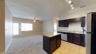 This single-floor residence showcases an open-concept layout that seamlessly connects the kitchen and living areas. The kitchen is outfitted with rich, dark espresso cabinetry that extends to the upper and lower walls, providing ample storage. A central island with a light-toned countertop offers additional workspace and casual seating potential. Stainless steel appliances, including a dishwasher and sink, add a modern, functional touch. Track lighting above the kitchen ensures focused illumination for food preparation. The transition from the kitchen's hardwood-style flooring to the plush carpeting of the living area creates a natural visual boundary between the two spaces. The living area benefits from a ceiling fan with an integrated light fixture, supporting both airflow and ambient lighting. Multiple windows throughout allow for natural light to permeate the space, enhancing the open and airy feel. Neutral-toned walls and a textured ceiling contribute to a clean, understated aesthetic throughout. A doorway visible toward the rear suggests access to additional rooms, adding to the overall functional layout of the home.