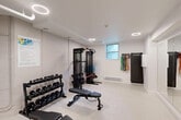This fitness facility at 5530 Ch De La Côte-Saint-Luc in Montréal, Quebec, offers a compact yet comprehensive exercise space designed for effective full-body workouts. The gym is thoughtfully laid out across a single floor, making efficient use of its footprint to accommodate a diverse selection of equipment.

The space is divided into distinct functional zones. The cardio area features multiple treadmills and a stationary bike, all positioned on dark rubber flooring that provides durability and traction. The strength training zone is well-stocked with a multi-functional cable machine, a tiered dumbbell rack with a full range of free weights, and adjustable workout benches.

Full-length mirrors line the walls, visually expanding the space and allowing users to monitor their form. The clean white walls and white flooring in the free weights area contrast with the dark rubber matting in the cardio zone, creating a modern, two-toned aesthetic. Circular halo-style recessed lighting illuminates the space evenly from the ceiling, contributing to a clean and contemporary look.

Climate control is provided by a wall-mounted air conditioning unit, ensuring comfort year-round. Small windows allow for a touch of natural light. The overall design is sleek, functional, and well-maintained, making it a practical fitness amenity.