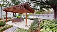 Located in the Peachtree Hills neighborhood of Atlanta, Georgia, this well-appointed outdoor communal space serves as a central gathering area for the residential community at 480 Peachtree Hills Ave NE. The grounds are defined by mature, towering oak trees that provide generous natural shade and a sense of established character throughout the courtyard.

A prominent wood pergola with a rich, warm stain anchors the space, positioned over a gravel-floored patio and paired with a curved brick knee wall that houses a built-in grilling station — ideal for outdoor entertaining. Nearby, a brick-edged fire pit area is furnished with bright blue Adirondack chairs, creating an inviting spot for relaxed gatherings.

Thoughtfully landscaped garden beds line brick-edged pathways that wind through the courtyard, filled with a diverse mix of shrubs, ground cover, ornamental grasses, and flowering plants including azaleas. The surrounding buildings feature classic painted brick exteriors with traditional window detailing, and select entries are accented with ornamental iron balconies and classic lantern-style lighting. The overall ambiance is tranquil and well-maintained, offering residents a verdant, park-like retreat in the heart of Atlanta.