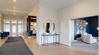 Located at 7405 Long Lake Rd in Columbus, OH, the Glendale Commons clubhouse is a thoughtfully designed single-story community space offering generous square footage across a well-appointed open layout. Visitors are welcomed by a bright, airy entrance corridor featuring light wood-look flooring, white structural columns, and a sleek console table topped with a round mirror — setting an immediately polished tone. The reception area houses a prominent front desk beneath cone-shaped pendant lights, accented by a textured feature wall and warm terracotta accent walls nearby. The kitchen and living area is a standout, boasting deep navy accent walls, grey shaker cabinetry, stainless steel appliances, and a long island with bar seating illuminated by a row of globe pendant lights. Adjacent lounge spaces offer versatile seating arrangements, including a sunlit sitting room with large windows, patterned drapery, and a curved sofa, as well as a dedicated media room with built-in seating and a wall-mounted display. Floor-to-ceiling glass doors at the rear flood the space with natural light and provide views of and access to an outdoor area. Recessed lighting throughout ensures a consistently bright and welcoming ambiance across all zones of this well-curated community clubhouse.