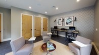 This single-floor amenity space is thoughtfully designed to serve as both a relaxed lounge and a functional business center. The interior features dark slate-tile flooring throughout, complemented by neutral-toned walls and recessed lighting that create a clean, contemporary atmosphere.

The lounge area is furnished with an eclectic mix of modern seating — including swivel chairs, accent chairs with ottomans, and side tables — arranged on a woven area rug. A large wall-mounted flat-screen television anchors one side of the room, while a wide window frames lush, tree-lined outdoor views and floods the space with natural light.

The business center area is equipped with a long wooden desk accommodating multiple computer workstations, ergonomic task chairs, and a shared printer. A textured, patterned accent wall provides a stylish backdrop, adorned with framed abstract artwork and industrial-style wall sconces that add a refined artistic touch.

Private rooms are accessible through warm wood-paneled doors with louvered vents, offering additional utility to the space. Large glass sliding doors on one end open to an outdoor area, enhancing the connection between interior and exterior. The overall design balances productivity and comfort, making it a versatile, well-appointed communal environment.