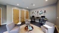 This single-floor amenity space is thoughtfully designed to serve as both a relaxed lounge and a functional business center. The interior features dark slate-tile flooring throughout, complemented by neutral-toned walls and recessed lighting that create a clean, contemporary atmosphere.

The lounge area is furnished with an eclectic mix of modern seating — including swivel chairs, accent chairs with ottomans, and side tables — arranged on a woven area rug. A large wall-mounted flat-screen television anchors one side of the room, while a wide window frames lush, tree-lined outdoor views and floods the space with natural light.

The business center area is equipped with a long wooden desk accommodating multiple computer workstations, ergonomic task chairs, and a shared printer. A textured, patterned accent wall provides a stylish backdrop, adorned with framed abstract artwork and industrial-style wall sconces that add a refined artistic touch.

Private rooms are accessible through warm wood-paneled doors with louvered vents, offering additional utility to the space. Large glass sliding doors on one end open to an outdoor area, enhancing the connection between interior and exterior. The overall design balances productivity and comfort, making it a versatile, well-appointed communal environment.