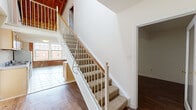Located on West Broad Street in Richmond, Virginia, this two-story unit blends industrial character with functional living spaces. The main level features warm hardwood flooring throughout the open living area, which flows directly into a galley-style kitchen equipped with honey-toned wood cabinetry, laminate countertops, a stainless steel sink, and white appliances including a range and refrigerator. Light tile flooring defines the kitchen and adjacent areas, providing a natural transition between zones. A carpeted staircase with a white metal and wood railing leads to the upper level, where exposed wooden beam ceilings and partial brick walls lend a distinctly industrial aesthetic. The loft-level landing opens to additional rooms with hardwood flooring, and a railing overlook connects the upper floor visually to the main living space below. The bathroom includes a vanity with wood cabinetry, a large mirror, tile flooring, and a full tub-shower combination. A secondary room on the lower level features rich dark hardwood floors and a glass-paneled door. Natural light filters in through multiple windows, and views of the surrounding streetscape add a sense of urban connectivity throughout the space.