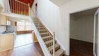 Located on West Broad Street in Richmond, Virginia, this two-story unit blends industrial character with functional living spaces. The main level features warm hardwood flooring throughout the open living area, which flows directly into a galley-style kitchen equipped with honey-toned wood cabinetry, laminate countertops, a stainless steel sink, and white appliances including a range and refrigerator. Light tile flooring defines the kitchen and adjacent areas, providing a natural transition between zones. A carpeted staircase with a white metal and wood railing leads to the upper level, where exposed wooden beam ceilings and partial brick walls lend a distinctly industrial aesthetic. The loft-level landing opens to additional rooms with hardwood flooring, and a railing overlook connects the upper floor visually to the main living space below. The bathroom includes a vanity with wood cabinetry, a large mirror, tile flooring, and a full tub-shower combination. A secondary room on the lower level features rich dark hardwood floors and a glass-paneled door. Natural light filters in through multiple windows, and views of the surrounding streetscape add a sense of urban connectivity throughout the space.