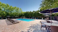 Located at 42 River Rd in Sunderland, Massachusetts, this outdoor pool area offers a well-appointed recreational retreat set within a beautifully landscaped environment. The centerpiece of the space is a large, rectangular swimming pool with crystal-clear blue water, designed to accommodate both leisurely swimming and relaxation. Depth markers are visible along the pool's edge, and entry is made convenient via stainless steel handrail ladders positioned at multiple points.

Surrounding the pool is a generously sized paved deck constructed with classic brick-patterned pavers, providing ample space for lounging and socializing. Rows of cushioned lounge chairs and chaise lounges line the perimeter of the pool, offering comfortable seating for sunbathers. Several shaded seating areas with large market umbrellas are also available, providing relief from the sun.

The pool area is fully enclosed by a dark metal security fence, ensuring safety and privacy. Mature, lush trees form a natural canopy around the entire space, creating a serene, park-like atmosphere while offering additional shade and seclusion. A brick clubhouse building with a pergola and additional outdoor seating areas is visible in the background, complementing the overall amenity space. The combination of open sky, vibrant greenery, and thoughtfully arranged furnishings makes this an inviting outdoor destination.