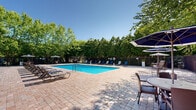 Located at 42 River Rd in Sunderland, Massachusetts, this outdoor pool area offers a well-appointed recreational retreat set within a beautifully landscaped environment. The centerpiece of the space is a large, rectangular swimming pool with crystal-clear blue water, designed to accommodate both leisurely swimming and relaxation. Depth markers are visible along the pool's edge, and entry is made convenient via stainless steel handrail ladders positioned at multiple points.

Surrounding the pool is a generously sized paved deck constructed with classic brick-patterned pavers, providing ample space for lounging and socializing. Rows of cushioned lounge chairs and chaise lounges line the perimeter of the pool, offering comfortable seating for sunbathers. Several shaded seating areas with large market umbrellas are also available, providing relief from the sun.

The pool area is fully enclosed by a dark metal security fence, ensuring safety and privacy. Mature, lush trees form a natural canopy around the entire space, creating a serene, park-like atmosphere while offering additional shade and seclusion. A brick clubhouse building with a pergola and additional outdoor seating areas is visible in the background, complementing the overall amenity space. The combination of open sky, vibrant greenery, and thoughtfully arranged furnishings makes this an inviting outdoor destination.