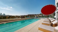 Located at 200 Mesnager Street in the heart of Los Angeles, this rooftop pool deck offers a striking outdoor amenity space defined by clean, modern architecture and open-sky exposure. The centerpiece is a generously sized lap pool with vivid turquoise water, framed by smooth light-toned tile decking that extends across the entire level. Adjacent to the main pool sits a separate spa, complete with accessibility equipment, ensuring the space is welcoming to all guests. A row of cushioned chaise lounges paired with bold red circular umbrellas lines one side of the pool, creating a vibrant, resort-style atmosphere ideal for relaxation under the Southern California sun. A shaded pergola structure adds a secondary lounge zone, complemented by potted greenery and planters that soften the hardscape. Glass perimeter railings preserve unobstructed panoramic views, showcasing the Downtown Los Angeles skyline to one side and the rolling hills of the surrounding neighborhood to the other. The multi-story building facade, finished in crisp white paneling with large windows and private balconies, rises directly alongside the deck, reinforcing the urban sophistication of the setting. The overall ambiance blends contemporary design with functional outdoor living, making this rooftop deck a distinctive and well-appointed amenity space.