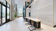This two-story space showcases a refined contemporary design defined by soaring double-height ceilings, floor-to-ceiling windows, and a carefully curated material palette. The ground level features large-format porcelain tile flooring throughout, anchored by a prominent reception area with a marble-topped desk set against an expansive marble-clad feature wall with gold trim detailing. Pendant light fixtures cascade dramatically from above, reinforcing the vertical scale of the space.

The open-plan layout includes multiple collaborative seating zones, a communal work table with gold-framed shelving units, and a built-in kitchenette with a white cabinetry bar tucked beneath the staircase. A sculptural open staircase with glass railings and dark metal handrails connects both levels, while elevator banks finished in brushed gold and mirrored panels provide additional vertical access.

Lounge areas are appointed with neutral upholstered sofas, marble coffee tables, and curated artwork including wall-mounted glass sculptures. Generous glazing on multiple facades floods the interior with natural light and provides views of the surrounding streetscape. Lush potted plants are positioned throughout, softening the clean architectural lines and adding a natural, welcoming element to the overall ambiance.