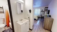 Located at 2500 University Blvd NE in Albuquerque, New Mexico, this compact yet thoughtfully arranged unit offers one bedroom and two bathrooms across a single floor. The space makes excellent use of its footprint, with wood-look plank flooring running throughout to create a cohesive and clean aesthetic.

The bedroom serves as the central living area and is immediately distinguished by a bold orange accent wall that adds warmth and character to the room. It is furnished with a full bed, a wooden dresser, a wall-mounted flat-screen television, a desk with seating, and open shelving that provides additional storage. Natural light filters in through a window fitted with vertical blinds.

A compact kitchenette is integrated into the layout, featuring white cabinetry, a microwave, a refrigerator, and a sink — offering convenient in-unit meal preparation.

The unit includes two bathrooms: one features a vanity with under-cabinet storage and a wall-mounted mirror, while the second contains a full bathtub and shower combination, a toilet, and overhead shelving for towels and toiletries. Both bathrooms are clean and functional in design.

A short entry corridor with light wood-look tile connects the front door to the main living space, providing a clear transition into the unit. Wall-mounted hair dryers in the bathroom area suggest a hospitality-oriented setup, making this unit well-suited for extended-stay or short-term accommodations.