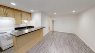 This single-floor space is designed with an open-concept layout that seamlessly connects the kitchen and living area, creating a functional and cohesive environment. The kitchen features a peninsula with a dark countertop, an undermount sink, and light wood cabinetry overhead, providing ample storage. The peninsula design allows for both food preparation and casual dining, while also maintaining a clear sightline into the adjoining living area.

The living space is generously proportioned and finished with light gray hardwood-style flooring that runs consistently throughout, contributing to a clean, unified aesthetic. Bright white walls are kept minimal and unadorned, allowing the space to feel open and airy. Recessed lighting is installed throughout the ceiling, providing even, modern illumination across both the kitchen and living areas.

A doorway visible from the main space leads to additional rooms, suggesting a functional layout beyond the open living zone. A wall-mounted radiator is also present, indicating a practical heating system integrated into the space. Overall, the design balances simplicity and functionality with contemporary finishes.