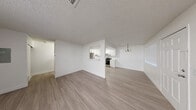 This single-floor unit offers a clean, functional layout with a bright and cohesive design throughout. The main living area is generously proportioned, featuring light wood-style luxury vinyl flooring, crisp white walls, and a textured ceiling that extends across the entire space. Natural light filters in through large windows fitted with horizontal blinds, creating a welcoming and airy atmosphere.

The living area flows seamlessly into the dining space, which is accented by a classic gold-toned chandelier. A half-wall with a pass-through window connects the dining area to the kitchen, offering an open feel while maintaining a defined layout. The kitchen is well-appointed with white shaker-style cabinetry, light countertops, a stainless steel refrigerator, a stainless steel electric range, an under-cabinet range hood, and a double-basin sink — all set against the same light flooring for a unified aesthetic.

The bedrooms continue the light and neutral palette, each offering ample space and sliding mirror-door closets that provide generous storage. One room also features an electrical panel, adding practical utility. A dedicated entry area leads to the front door, completing the thoughtful and efficient use of space throughout this well-maintained unit.