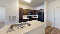 Located at 2913 Hunter Rd in San Marcos, TX, this single-floor, 2-bedroom, 2-bathroom residence offers a well-organized layout designed for comfortable everyday living. The kitchen serves as the heart of the home, featuring rich dark wood cabinetry extending to the upper walls, light countertops, and a full suite of black appliances including a range, microwave, and refrigerator. A central island with a built-in sink and breakfast bar connects the kitchen to the living area, creating an open, functional flow throughout. Wood-style flooring runs throughout the main living and kitchen areas, adding warmth and visual continuity. Both bedrooms are carpeted and bathed in natural light from windows with blinds, each equipped with ceiling fans. The primary bedroom is a comfortable size and adjoins a private en-suite bathroom complete with a dual-sink vanity featuring dark cabinetry, a tub-shower combo, and direct access to a walk-in closet. The second bathroom mirrors the same design aesthetic with a single-sink vanity and tub-shower combination. A hallway and dedicated closet provide additional storage throughout the home, rounding out a practical and cohesive living space.