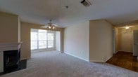 This single-story space presents a spacious, open living area finished with wall-to-wall neutral-toned carpeting that lends a comfortable, understated feel underfoot. The room is anchored by a ceiling fan with integrated lighting, providing both air circulation and ambient illumination. A prominent arched window along one wall allows natural light to flood the interior, creating a bright and welcoming atmosphere. The walls are painted in soft, warm beige tones that complement the light carpeting and contribute to the room's airy quality. A textured popcorn ceiling adds a classic residential detail overhead, while white baseboards provide clean definition along the floor line. A central HVAC vent is positioned in the ceiling, ensuring efficient climate control throughout the space. The open layout offers flexible potential for various furniture arrangements, making it adaptable for a range of uses. Overall, the space maintains a clean, neutral palette that serves as a versatile backdrop, ready to accommodate personal décor preferences.