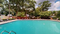 Located at 5000 S Lincoln Trace Ave SE in Smyrna, Georgia, this outdoor pool facility offers a well-appointed recreational retreat set against a backdrop of mature, towering trees. The centerpiece is a generously sized aquamarine swimming pool with clearly marked shallow and deep ends, accessible via stainless steel entry rails. The pool is divided by a safety rope, and the surrounding deck is finished in a clean, neutral tile that complements the water's vivid color.

The pool deck extends broadly on multiple sides, providing ample space for lounging and socializing. Rows of sleek chaise lounge chairs line the perimeter, ideal for relaxing in the open air. A dedicated patio area with umbrella-shaded seating offers a comfortable spot for shade and gathering. A charming structure with a distinctive conical roof serves as a focal point on one end of the deck, adding architectural character to the space.

The entire area is enclosed by a black iron fence with brick accent sections, ensuring both safety and a polished aesthetic. Lush greenery and mature deciduous trees frame the space on all sides, creating a sense of natural seclusion while allowing abundant natural light to flood the area. The overall ambiance is inviting, resort-like, and well-suited for warm-weather recreation.