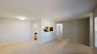 This single-story, one-bedroom, one-bathroom unit offers a practical and efficient layout designed to maximize usable space. The open living area is finished with neutral-toned carpeting and light gray walls, creating a clean and versatile backdrop. Two windows allow natural light to fill the room, providing views of the surrounding exterior.

The kitchen is positioned adjacent to the living area and features a pass-through counter opening, enhancing the sense of connectivity between spaces. Inside, the kitchen is equipped with white cabinetry, a refrigerator, stove, microwave, and dishwasher. Light wood-style flooring runs throughout the kitchen, complementing the crisp cabinetry. A tiled backsplash adds a subtle decorative element.

A dedicated hallway connects the main living area to the bedroom, bathroom, and a convenient in-unit laundry closet housing a stacked washer and dryer. The bedroom is generously proportioned with soft carpeting and two windows that bring in natural light. The bathroom features a full tub and shower combination with white tile surround. The overall design is clean, functional, and well-suited for comfortable everyday living.