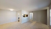 This single-story, one-bedroom, one-bathroom unit offers a practical and efficient layout designed to maximize usable space. The open living area is finished with neutral-toned carpeting and light gray walls, creating a clean and versatile backdrop. Two windows allow natural light to fill the room, providing views of the surrounding exterior.

The kitchen is positioned adjacent to the living area and features a pass-through counter opening, enhancing the sense of connectivity between spaces. Inside, the kitchen is equipped with white cabinetry, a refrigerator, stove, microwave, and dishwasher. Light wood-style flooring runs throughout the kitchen, complementing the crisp cabinetry. A tiled backsplash adds a subtle decorative element.

A dedicated hallway connects the main living area to the bedroom, bathroom, and a convenient in-unit laundry closet housing a stacked washer and dryer. The bedroom is generously proportioned with soft carpeting and two windows that bring in natural light. The bathroom features a full tub and shower combination with white tile surround. The overall design is clean, functional, and well-suited for comfortable everyday living.