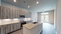 This single-story residence in Houston, TX offers a thoughtfully designed open-concept layout that seamlessly connects the kitchen and living areas. The kitchen is a standout feature, showcasing wood-grain cabinetry in a warm grey tone, light quartz countertops, and a full suite of stainless steel appliances including a French door refrigerator, electric range, and built-in microwave. A central island with an undermount sink and gooseneck faucet provides additional prep space and casual seating potential. A white subway tile backsplash adds a clean, modern contrast to the cabinetry. Wood-look flooring extends throughout the main living areas, contributing to a cohesive and contemporary aesthetic. Recessed lighting illuminates the space efficiently throughout. The living area benefits from natural light through French doors topped with a transom window, which open onto a private balcony with outdoor views. A ceiling fan overhead enhances comfort. The bathroom features large-format tile flooring, a wood-grain vanity with a light countertop, and a walk-in shower. The bedroom is carpeted and connects conveniently to the bathroom, completing this well-appointed Houston residence.