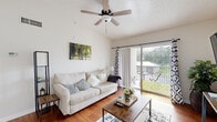 Located at 3717–3719 SW Coquina Cove Way in Palm City, Florida, this single-floor residence offers a well-organized layout designed for comfortable everyday living. The bright living room is a standout space, featuring vaulted ceilings, warm hardwood flooring, and a ceiling fan overhead. Large sliding glass doors flood the room with natural light and open directly onto a private balcony with outdoor seating, offering pleasant views of the surrounding greenery. A dedicated dining area sits adjacent to the living space, highlighted by hardwood floors, a large window with vertical blinds, and ample room for a four-person dining set. The kitchen is outfitted with white cabinetry, white appliances including a refrigerator and electric range, and generous counter space. A convenient in-unit laundry area is accessible directly from the kitchen. The primary bedroom features vaulted ceilings, a ceiling fan, carpeted floors, and dual closets, while the second bedroom is quiet and well-lit with carpeted flooring. The bathroom is clean and functional, complete with a tub-shower combination, a single-sink vanity with ample cabinet storage, and tile flooring. Throughout the residence, a neutral color palette and natural light contribute to an airy, welcoming atmosphere.
