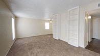 This single-floor space presents a clean, functional design centered around an open living area finished in warm neutral tones. The room is carpeted throughout in a beige, low-pile material that adds softness underfoot and contributes to the space's understated, comfortable feel. The walls are painted in a light cream tone, creating a bright and airy atmosphere that complements the natural light entering through a centrally positioned window.

The ceiling features a classic textured finish and is equipped with a ceiling fan, providing both practical ventilation and a modest decorative element. A notable architectural detail is the pair of tall, white built-in shelving units positioned along one wall. These floor-to-ceiling open shelves offer generous display and storage capacity, adding structure and visual interest to the room while maintaining the overall neutral palette.

The layout is open and unobstructed, lending flexibility for a variety of furniture arrangements and uses. White baseboards frame the perimeter of the room, adding a clean finishing detail. Overall, the space conveys a straightforward, well-maintained character suited to a range of functional needs.