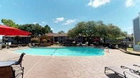 This outdoor amenity area centers around a large, freeform swimming pool with clear turquoise water, set within a spacious stamped concrete deck designed to accommodate ample lounge chairs and poolside seating. The pool is enclosed by a wrought iron fence, providing both security and an open visual connection to the surrounding landscaped grounds and mature shade trees.

A standout feature of the space is a substantial wooden pergola structure, constructed with natural timber framing and positioned adjacent to the pool deck. Beneath it sits a built-in outdoor kitchen with stone masonry detailing and a granite-style countertop, making it well-suited for outdoor gatherings. The pergola's slatted roof casts patterned shadows across the stamped concrete patio below.

A single-story poolside building borders the deck, featuring French doors, large windows with horizontal blinds, and a covered porch area with seating. Potted flowering plants accent the entryway, adding a welcoming touch. The building appears to house a fitness facility, visible through the windows. The overall environment benefits from abundant natural sunlight and the shade of mature oak trees, creating a comfortable and well-appointed outdoor retreat.