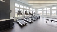 Located at 1600 Lake Seymour Dr in Middletown, Delaware, this well-appointed fitness center offers a comprehensive workout environment designed to accommodate a wide range of exercise routines. The single-floor facility is characterized by soaring ceilings and an abundance of natural light, courtesy of expansive floor-to-ceiling windows that line multiple walls, offering views of the surrounding community and a poolside area.

The cardio zone features a row of treadmills and elliptical machines, all equipped with individual screens and positioned directly in front of large windows to maximize outdoor views during workouts. A built-in cubby storage unit nearby provides convenient space for personal belongings.

The strength and functional training area is well-stocked with a full rack of dumbbells, kettlebells of varying weights, medicine balls, stability balls, and a cable cross-trainer machine — catering to both beginners and experienced athletes. Large mirrors are strategically placed to assist with form and to visually expand the space.

The flooring throughout consists of durable speckled rubber, ideal for a high-traffic fitness environment. The interior design features a clean, modern aesthetic with a two-tone color scheme of crisp white and deep slate blue accent walls, complemented by recessed and linear lighting. An on-demand fitness screen is also present, supporting guided workout sessions.