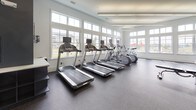Located at 1600 Lake Seymour Dr in Middletown, Delaware, this well-appointed fitness center offers a comprehensive workout environment designed to accommodate a wide range of exercise routines. The single-floor facility is characterized by soaring ceilings and an abundance of natural light, courtesy of expansive floor-to-ceiling windows that line multiple walls, offering views of the surrounding community and a poolside area.

The cardio zone features a row of treadmills and elliptical machines, all equipped with individual screens and positioned directly in front of large windows to maximize outdoor views during workouts. A built-in cubby storage unit nearby provides convenient space for personal belongings.

The strength and functional training area is well-stocked with a full rack of dumbbells, kettlebells of varying weights, medicine balls, stability balls, and a cable cross-trainer machine — catering to both beginners and experienced athletes. Large mirrors are strategically placed to assist with form and to visually expand the space.

The flooring throughout consists of durable speckled rubber, ideal for a high-traffic fitness environment. The interior design features a clean, modern aesthetic with a two-tone color scheme of crisp white and deep slate blue accent walls, complemented by recessed and linear lighting. An on-demand fitness screen is also present, supporting guided workout sessions.