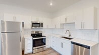 This thoughtfully designed one-bedroom, one-bathroom unit offers a clean, modern aesthetic across its single floor. The open kitchen is a standout feature, showcasing white flat-front cabinetry, a crisp subway tile backsplash, white quartz countertops, and a full suite of stainless steel appliances including a refrigerator, electric range, over-the-range microwave, and dishwasher. Wood-look plank flooring runs throughout the main living areas, lending warmth and continuity to the space. A bright hallway connects the rooms, with a glass-paneled door at the end drawing in natural light and offering a view of surrounding greenery. The bedroom is a comfortable, well-proportioned retreat finished with plush neutral carpeting and a window fitted with blinds that frames an outdoor tree view. A dedicated walk-in closet adjacent to the bedroom provides organized storage with a full-width hanging rod. The bathroom continues the unit's clean design language, featuring a white vanity with an undermount sink, a framed mirror, brushed nickel fixtures, and a full shower-tub combination. Recessed lighting throughout and crisp white walls give the entire unit a bright, airy feel that maximizes the sense of space.