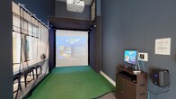 This single-floor space combines a sophisticated lounge area with a dedicated multi-sport simulator bay, creating a versatile amenity designed for both leisure and active entertainment. The lounge portion of the space is defined by soaring double-height ceilings and large windows that invite abundant natural light, framing views of outdoor greenery. The seating area is thoughtfully arranged with a plush brown sofa, striped accent chairs, and a round wood-and-metal coffee table, all set atop neutral carpeting. Elegant pendant lighting and a tall floor lamp contribute to the warm, inviting ambiance, while a translucent decorative partition adds architectural interest.

Adjacent to the lounge, the simulator bay transitions dramatically in aesthetic, with dark navy walls enclosing a dedicated turf-floored hitting area. A ceiling-mounted projector displays multi-sport simulation software onto a large impact screen framed by a sturdy net enclosure. Track lighting lines the top of the simulator structure, and a dedicated computer workstation beside the bay allows users to select from various sports and settings. The combination of an airy, light-filled social lounge and a sleek, immersive simulator environment makes this space a compelling and well-rounded amenity.