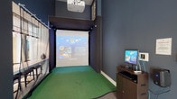 This single-floor space combines a sophisticated lounge area with a dedicated multi-sport simulator bay, creating a versatile amenity designed for both leisure and active entertainment. The lounge portion of the space is defined by soaring double-height ceilings and large windows that invite abundant natural light, framing views of outdoor greenery. The seating area is thoughtfully arranged with a plush brown sofa, striped accent chairs, and a round wood-and-metal coffee table, all set atop neutral carpeting. Elegant pendant lighting and a tall floor lamp contribute to the warm, inviting ambiance, while a translucent decorative partition adds architectural interest.

Adjacent to the lounge, the simulator bay transitions dramatically in aesthetic, with dark navy walls enclosing a dedicated turf-floored hitting area. A ceiling-mounted projector displays multi-sport simulation software onto a large impact screen framed by a sturdy net enclosure. Track lighting lines the top of the simulator structure, and a dedicated computer workstation beside the bay allows users to select from various sports and settings. The combination of an airy, light-filled social lounge and a sleek, immersive simulator environment makes this space a compelling and well-rounded amenity.