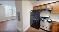 Unit 215 at 1221 E Brookland Park Blvd in Richmond, VA is a compact and efficiently designed 1-bedroom, 2-bathroom apartment spanning 400 square feet on a single floor. Warm-toned hardwood-style flooring runs throughout the living and bedroom areas, providing a cohesive and inviting aesthetic. The 120-square-foot bedroom features neutral beige walls, bright natural light from a window with blinds offering outdoor views of greenery, and clean white baseboards. The kitchen is well-appointed with warm wood cabinetry, granite-style countertops, stainless steel range, black refrigerator, and a double sink — maximizing functionality within a compact footprint. The unit includes two bathrooms: one featuring a vanity with wood cabinetry, a wall mirror, and light tile flooring, and a second with an accessible walk-in shower stall equipped with stainless steel grab bars and a handheld showerhead, making it suitable for accessibility needs. The open-concept layout, visible in the dollhouse view, integrates the living area and kitchen seamlessly via an archway. Neutral wall tones and consistent finishes throughout contribute to a clean, move-in-ready atmosphere.