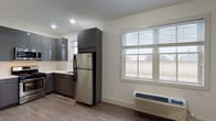Located at Village Place in Bay Shore, NY, this single-floor unit offers a clean, modern design throughout. The kitchen is outfitted with high-gloss dark gray cabinetry, stainless steel appliances including a full-size refrigerator, gas range, and over-the-range microwave, complemented by light countertops and recessed lighting. A window over the kitchen area brings in natural light, while a wall-mounted HVAC unit ensures year-round comfort. The open living area flows naturally from the kitchen, featuring wood-look luxury vinyl plank flooring that extends throughout the entire unit, creating a cohesive and contemporary aesthetic. Bright white walls and neutral tones keep the space feeling clean and airy. The bedroom is a well-proportioned, private retreat accessed through a panel-style door, complete with a ceiling fan and a built-in closet with dual hanging rods for ample storage. The bathroom is finished with light tile flooring, a sleek vanity with a white countertop, and a full tub and shower enclosure featuring frameless sliding glass doors. Every detail of this unit reflects a thoughtfully modern and functional design, well-suited for comfortable everyday living in the heart of Bay Shore.