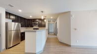 This well-designed single-floor residence offers a clean, contemporary aesthetic carried consistently throughout its layout. The kitchen serves as a functional centerpiece, showcasing rich dark cabinetry, light granite countertops, and a full suite of stainless steel appliances including a refrigerator, range, and microwave. A peninsula with pendant lighting overhead provides additional prep space and creates a natural transition into the open living area. Light hardwood-style flooring runs throughout the entire unit, contributing to a cohesive and airy feel.

The bedrooms are generously proportioned, each finished with the same light flooring and neutral wall tones. One bedroom features a ceiling fan and a window fitted with blinds, allowing for natural light and comfortable airflow. Additional bedrooms offer similarly bright, open layouts with windowed walls and wide doorways. The unit includes two full bathrooms, each outfitted with a tub and shower combination, chrome fixtures, and clean white surrounds that complement the overall minimalist palette.

A central hallway connects the living spaces efficiently, and the floorplan—as seen in the dollhouse overview—offers a thoughtful, functional arrangement. The consistent use of neutral tones, modern finishes, and natural light gives the entire space a fresh, move-in-ready character.