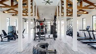 Located at 4711 Azalea Landing Dr in Wilmington, NC, this impressive fitness center delivers a high-end workout experience within a generously sized, single-floor layout. The space is defined by its dramatic vaulted ceilings adorned with exposed rustic wood beam trusses, which lend a warm, architectural character that contrasts beautifully with the crisp white walls and structural columns throughout. Large black-framed windows line the perimeter, flooding the interior with natural light and offering views of the surrounding outdoor areas.

The flooring features durable light grey wood-look planks that flow seamlessly across the entire space. A standout design element is the bold, wave-patterned accent wall near the entrance, adding an artistic focal point to the room. Overhead, industrial-style ceiling fans and pendant lighting ensure comfort and visibility throughout.

Equipment is thoughtfully arranged to accommodate a wide range of workouts, including a power rack with free weights and barbells, cable machines, a heavy punching bag, rowing machines, treadmills, and cardio equipment. A well-stocked wall-mounted rack holds medicine balls, kettlebells, and resistance tools. A large garage-style roll-up door opens directly to an outdoor lawn area, seamlessly blending indoor and outdoor fitness capabilities.