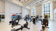 Located at 2300 N Lincoln Park West in Chicago's Lincoln Park neighborhood, this two-story fitness center is a thoughtfully designed facility that blends architectural elegance with comprehensive workout amenities. The main floor is defined by soaring double-height ceilings, light hardwood flooring, and dramatic floor-to-ceiling arched windows that flood the space with natural light and offer serene views of the surrounding greenery. Globe pendant chandeliers add a refined touch overhead, while an array of cardio equipment — including ICG cycling bikes, rowing machines, treadmills, and ellipticals — is arranged throughout the open layout. A decorative staircase with ornate metalwork railings connects the two levels, serving as a striking architectural focal point. A glass-enclosed studio space is accessible from the main floor, providing a dedicated area for group or individual programming. The upper level features a strength-focused zone with rubber flooring, free weights, dumbbell racks, cable machines, a squat rack, and various resistance equipment. Floor-to-ceiling mirrors and linear recessed lighting give this level a clean, modern aesthetic, while arched windows continue to bring in natural light. Together, the two floors offer a well-rounded fitness environment that is both functional and refined.