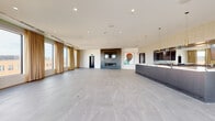 Located at 4001 Dr. Martin Luther King Jr. Blvd in Kansas City, Missouri, this single-floor space offers a contemporary, open-plan layout designed for versatile use. Large-format gray porcelain tile flooring extends throughout, lending a clean, cohesive aesthetic to the expansive interior.

A striking linear fireplace set within a dark, recessed surround serves as a central focal point, accompanied by a mounted display screen above. Adjacent to the fireplace, a bold portrait artwork adds a distinct cultural touch to the otherwise minimalist design. Elevator access is conveniently positioned nearby.

A full-length bar and service counter anchors one side of the space, featuring fluted cabinetry in a cool gray tone, a mirrored back wall, stainless steel fixtures, and pendant lighting with brass accents. This area is further illuminated by dual mounted televisions and recessed ceiling lighting throughout.

A generous bank of floor-to-ceiling windows dressed with warm gold drapery lines the perimeter, flooding the interior with natural light and offering views of the surrounding Kansas City neighborhood. A glass sliding door opens to an outdoor terrace, extending the usable space further. The overall ambiance is polished, modern, and well-suited for events or gatherings.