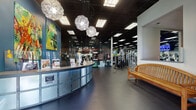 This comprehensive fitness facility is thoughtfully designed to accommodate a wide range of workout disciplines across a single expansive floor. Members are welcomed by a curved reception desk accented with distinctive globe pendant lighting, teal feature walls, and colorful artwork that set an energetic yet inviting tone throughout the space. The main workout floor stretches generously, featuring durable rubber flooring underfoot and a dark drop ceiling with grid lighting overhead, housing a broad selection of cardio machines — including treadmills, stationary bikes, and ellipticals — alongside a well-equipped strength training zone with cable machines and selectorized equipment. Natural light filters in through large windows, brightening the cardio and stretching areas. A dedicated spinning studio provides an immersive cycling experience, complete with projection screens, dynamic lighting effects, and rows of professional spinning bikes. A separate multi-purpose studio offers a large projection screen, TRX suspension trainers, kettle bells, and medicine balls for group classes and personal training sessions. Glass partition doors separate functional zones, enhancing both visibility and flow. The facility also includes a full-size indoor basketball court with a polished hardwood floor, painted court lines, retractable hoops, high ceilings, and protective wall padding — ideal for team sports and recreational play.