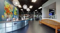 This comprehensive fitness facility is thoughtfully designed to accommodate a wide range of workout disciplines across a single expansive floor. Members are welcomed by a curved reception desk accented with distinctive globe pendant lighting, teal feature walls, and colorful artwork that set an energetic yet inviting tone throughout the space. The main workout floor stretches generously, featuring durable rubber flooring underfoot and a dark drop ceiling with grid lighting overhead, housing a broad selection of cardio machines — including treadmills, stationary bikes, and ellipticals — alongside a well-equipped strength training zone with cable machines and selectorized equipment. Natural light filters in through large windows, brightening the cardio and stretching areas. A dedicated spinning studio provides an immersive cycling experience, complete with projection screens, dynamic lighting effects, and rows of professional spinning bikes. A separate multi-purpose studio offers a large projection screen, TRX suspension trainers, kettle bells, and medicine balls for group classes and personal training sessions. Glass partition doors separate functional zones, enhancing both visibility and flow. The facility also includes a full-size indoor basketball court with a polished hardwood floor, painted court lines, retractable hoops, high ceilings, and protective wall padding — ideal for team sports and recreational play.