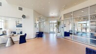 This contemporary two-story amenity space is designed with a clean, modern aesthetic throughout, characterized by crisp white walls, recessed lighting, and an open, airy atmosphere. The main level greets visitors with a spacious lobby featuring warm tile flooring, floor-to-ceiling glass entry doors, and a glass-partitioned office or leasing area. Generous seating with distinctive navy blue velvet chairs provides comfortable gathering spots, while potted tall plants add organic accents to the bright, welcoming interior. An adjacent lounge area offers a sophisticated retreat complete with a sleek linear fireplace, wall-mounted television, built-in kitchen counter with bar-height seating in warm leather tones, and expansive floor-to-ceiling windows that flood the space with natural light and provide direct access to the outdoor terrace. The fitness center is well-equipped with exercise machines, a wall-mounted television, a water station, and rubber flooring, offering direct outdoor access. A dedicated game room features a foosball table beneath pendant lighting, framed artwork, and a playful atmosphere. The second floor hosts additional lounge seating on plush patterned carpet alongside vibrant abstract artwork, with bright windows overlooking the surrounding skyline. The crowning feature is a spacious covered rooftop terrace offering expansive views of the mountains and surrounding community, furnished with multiple sectional seating arrangements and a ceiling fan for year-round enjoyment.