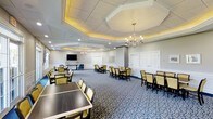 This refined event space presents an impressive interior defined by its striking architectural ceiling design. A large octagonal coffered tray ceiling serves as the room's focal point, accented by warm ambient uplighting that highlights the layered crown molding details. Two classic candelabra-style chandeliers provide additional illumination, complementing the recessed lighting throughout.

The generously sized room accommodates multiple dining tables arranged to maximize seating capacity, each surrounded by cushioned yellow-green upholstered chairs set against dark wood table frames. The flooring features a bold navy blue patterned carpet with scrolling motifs, adding visual richness underfoot.

Soft grey walls with wainscoting paneling lend a traditional, formal character to the space, while white paneled doors and trim provide clean contrast. A wall-mounted flat-screen television is installed at one end, supporting presentations or entertainment during events. A small accent console table with decorative greenery adds a refined finishing touch. The overall ambiance is formal yet welcoming, making this space well-suited for banquets, receptions, or group dining occasions.