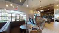 This single-floor clubroom and media room in Cypress, TX, offers a thoughtfully designed communal space that balances social gathering with relaxed lounge comfort. The open layout is anchored by a formal dining area seating up to ten, furnished with plush velvet chairs surrounding a solid wood table and crowned by a striking brass and glass globe chandelier. Pendant lighting throughout reinforces a warm, upscale ambiance. Adjacent to the dining space, a full wet bar features sleek dark cabinetry, a quartz-topped island, a built-in sink, and a dramatic dark hexagonal tile accent wall — all illuminated by brushed gold pendant lights and a wall-mounted display screen. The lounge area is furnished with rich leather sofas, a tufted ottoman, and woven rattan accent chairs, arranged atop layered area rugs on light hardwood-style flooring. A curated collection of large-scale abstract artwork adorns the walls throughout, adding an artistic character to the space. Oversized windows and glass doors invite abundant natural light and outdoor views of the surrounding palm-lined community. The media room area offers an intimate seating arrangement ideal for private viewing or casual gatherings, completing this well-appointed community amenity space.