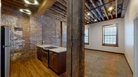 Located at 417 S Second St in Leavenworth, Kansas, this single-floor space is a compelling example of historic adaptive reuse, where original industrial character has been thoughtfully preserved alongside modern updates. The building's most striking architectural feature is its exposed ceiling structure, where heavy timber beams and wood planking span the full width of the space, lending a raw, warehouse-like aesthetic that sets the tone throughout.

The kitchen is a standout area, featuring rich dark wood cabinetry, granite-look countertops, and stainless steel appliances including a range and refrigerator. Original exposed brick walls enclose the kitchen on multiple sides, reinforcing the building's historic identity. A substantial structural post in reclaimed wood anchors the transition between the kitchen and the adjacent open living area, adding to the space's authentic character.

The open carpeted room beyond the kitchen offers generous proportions, with two wood-trimmed windows that bring in natural light. Exposed brick continues along one wall, while track lighting mounted to the timber ceiling provides functional illumination. A separate, carpeted room with its own window and exposed beam ceiling offers additional flexible space. The bathroom is cleanly finished with wood-look flooring, tile surround, and a tub-shower combination, providing a modern contrast to the building's historic bones.