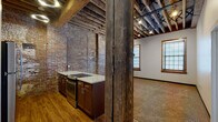 Located at 417 S Second St in Leavenworth, Kansas, this single-floor space is a compelling example of historic adaptive reuse, where original industrial character has been thoughtfully preserved alongside modern updates. The building's most striking architectural feature is its exposed ceiling structure, where heavy timber beams and wood planking span the full width of the space, lending a raw, warehouse-like aesthetic that sets the tone throughout.

The kitchen is a standout area, featuring rich dark wood cabinetry, granite-look countertops, and stainless steel appliances including a range and refrigerator. Original exposed brick walls enclose the kitchen on multiple sides, reinforcing the building's historic identity. A substantial structural post in reclaimed wood anchors the transition between the kitchen and the adjacent open living area, adding to the space's authentic character.

The open carpeted room beyond the kitchen offers generous proportions, with two wood-trimmed windows that bring in natural light. Exposed brick continues along one wall, while track lighting mounted to the timber ceiling provides functional illumination. A separate, carpeted room with its own window and exposed beam ceiling offers additional flexible space. The bathroom is cleanly finished with wood-look flooring, tile surround, and a tub-shower combination, providing a modern contrast to the building's historic bones.