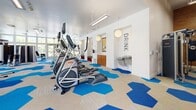 This Fitness Center & Outdoor Game Zone is a comprehensive recreational facility spread across a single floor, offering a wide range of amenities for both indoor and outdoor activity. The main gym area is bright and expansive, lined with floor-to-ceiling windows that flood the space with natural light and provide views of the surrounding greenery. The gym is outfitted with rows of cardio machines, including ellipticals and stationary bikes, as well as cable machines and free weights. A distinctive hexagonal patterned carpet in bold blue and neutral tones runs throughout, adding a vibrant, modern aesthetic. A dedicated strength and studio room features warm wood-paneled walls, full-length mirrors, pop-art wall décor, exercise benches, medicine balls, and a workout mat — creating an energetic environment for functional training. The facility also includes a game room, two bathrooms, a closet, and additional support spaces. The entrance area is light-filled with large windows and glass double doors opening to the outdoors. Outside, well-maintained sport courts — suitable for tennis and basketball — are surrounded by thoughtfully landscaped gardens with mature trees, colorful shrubs, and manicured pathways, all set beneath open blue skies.