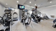 This fitness center offers a comprehensive, single-floor workout facility designed to support a wide range of cardio-focused exercise routines. The space is thoughtfully laid out with a generous open floor plan that accommodates an extensive selection of equipment, including treadmills, StairMaster steppers, elliptical trainers, upright and recumbent stationary bikes, and a rowing machine, among other machines by brands such as Cybex.

The interior features clean white walls, recessed ceiling lighting, and durable carpeted flooring that spans the entire space. Multiple wall-mounted flat-screen televisions are positioned throughout the room, allowing users to stay entertained during their workouts. A built-in water fountain is conveniently located within the facility for easy access.

Natural light filters into the space through several windows, offering views of lush tropical landscaping with palm trees and neighboring rooftops, creating a bright and inviting atmosphere. Ceiling-mounted speakers suggest an integrated sound system for an enhanced workout experience.

The fitness center is accessible via a glass-paneled entry door and includes clearly marked emergency exits, a fire extinguisher, and proper ventilation through visible HVAC systems. The overall ambiance is clean, modern, and functional, making it a well-appointed destination for daily fitness routines.